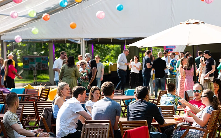 A crowd of people seated under giant umbrellas, next to a marquee for a corporate summer party company offsites
