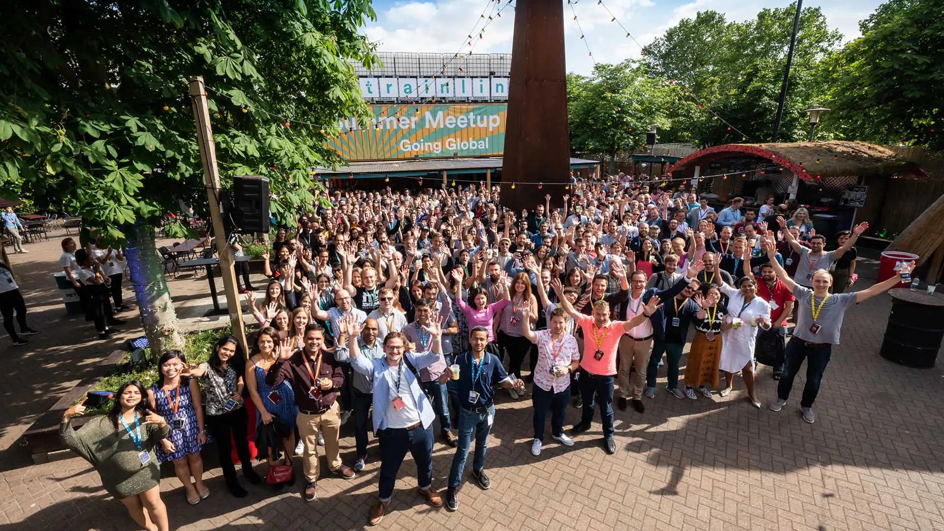 A huge crowd of people standing outside, in the sunshine, with arms raised, waving at the camera for a team photo at a team away day event