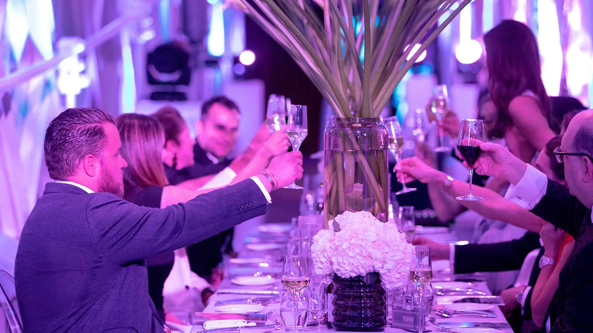 People in business suits sitting at a long dining table, raising glasses of champagne in a toast at a corporate event