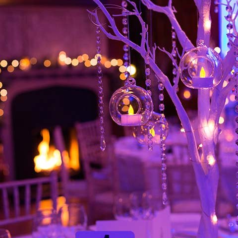 Tables with bare white trees decorated with see through Christmas baubles and strings of glass beads sit in front of a roaring open fire, an LED lighted Christmas garland sits above the fireplace creating a Corporate Christmas Party Atmosphere