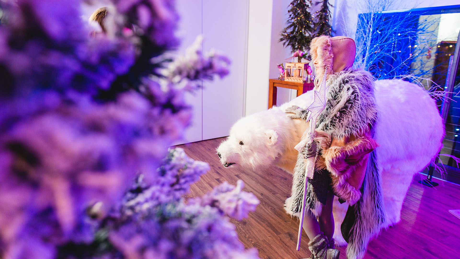 An out of focus Christmas tree with a woman wearing a fur cloak, walking with a Polar bear