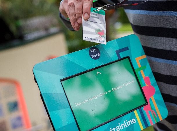 A badge is about to be tapped against a “Tap here” section on the screen of a kiosk, which is part of an interactive display at an event