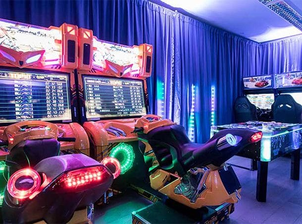 An image of an amusement arcade set up at a private home, two motorbike arcade games with scores on the board above. Blue lighting.