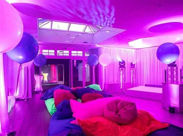 A cosy room with blue and pink balloons, pink-purple lighting, and bean bags on the floor, ready for a party.