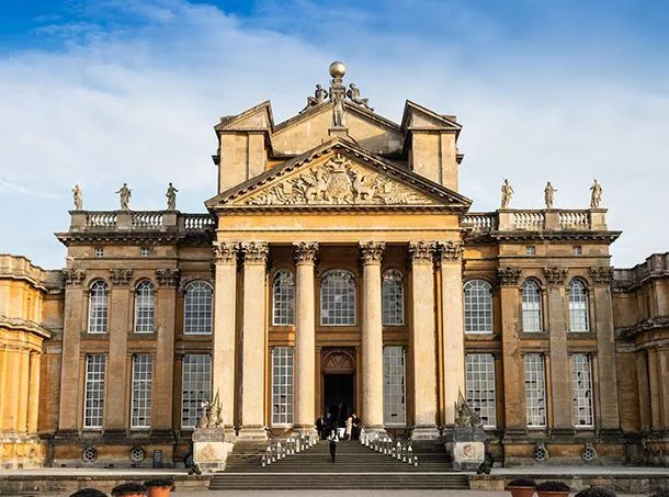 An image of Blenheim Palace. A large historical building with columns, long arched windows, and a wide set of stairs leading up to the entrance.