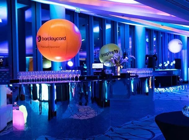 An indoor bar setting with blue and purple lighting creating a cool atmosphere. Balloons with brand names on them above rows of wine glasses.