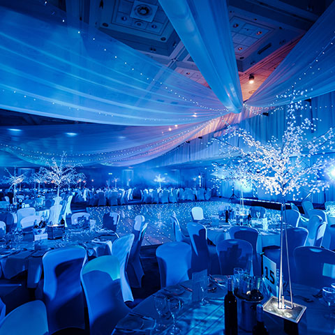An image of a Christmas event. Multiple tables are set up with a dance floor at the front, with lots of blue twinkling lights for a wintery atmosphere.