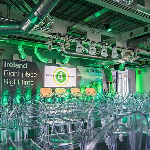 A modern, industrial event venue set up for a conference, with a large screen, stage, seating and lectern for speakers and seating laid out for delegates