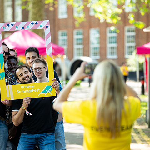 summer party venue london An outdoor setting. A woman is taking a photo of a group of men with their faces inside a physical frame that reads 'SummerFest'.