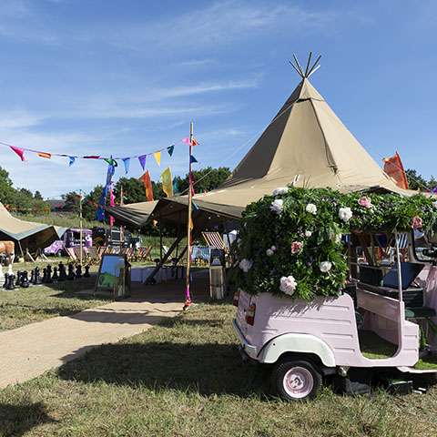 Summer party organisers MGN events An outdoor image of a tipi marquee, a small light pink vehicle covered in flowers, and colourful bunting. All in the sunshine on the grass.