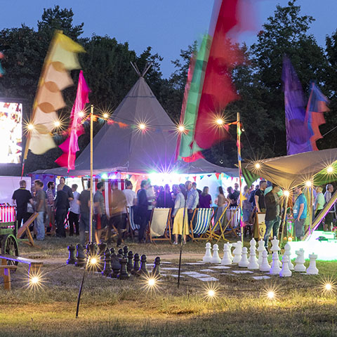Family fun days A Tipi marquee surrounded by festival flags, lighting and party-goers and with a giant chess set in the foreground, in the early evening