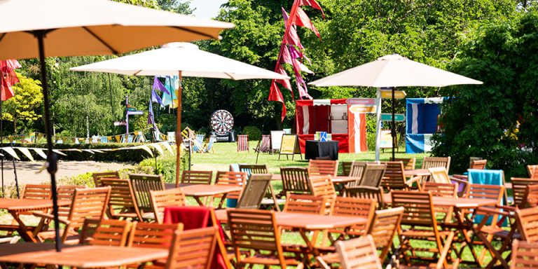 Corporate festivals An outdoor party set-up featuring wooden tables and chairs, some umbrellas, and party games in the distance. A backdrop of trees.