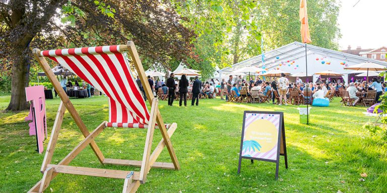 Company festivals landscape image of a summer event. At the front is a red and white striped sun lounger facing a crowded area with a white marquee.