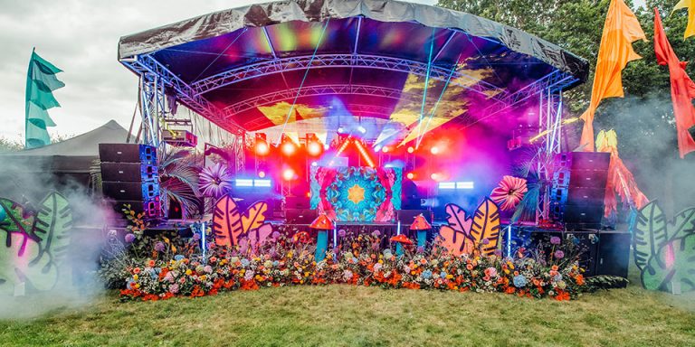 A large stage decorated with psychedelic coloured decorations and lighting, giant toadstools, flowers and flags, with dry ice being released to each side of the stage