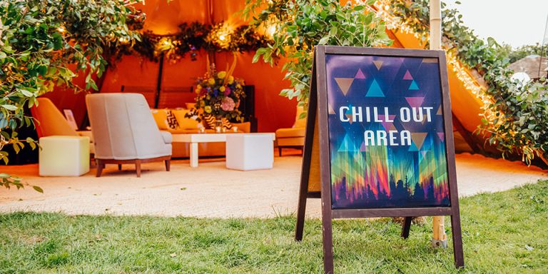 An a frame sign says 'Chill Out Area', in the background, partially out of focus, is a TP marquee with festoon lighting, furniture and floral decoration