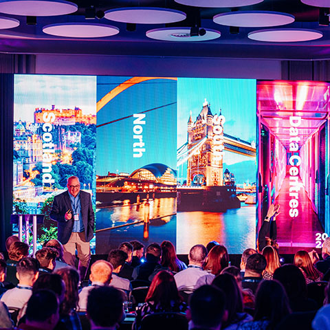 A man stand in front of a crowd of seated delegates as he presents at a company kick-off event. The screen behind him is split into 4 segments labelled, Scotland, North, South and data centre