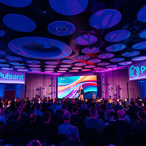 A crowd of seated delegates with a group of people facing them, seated on stage in front of a large presentation screen, above them a dark ceiling is decorated with circular lighting effects