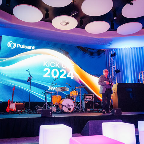 A man stands on a stage, beside him is a set of drums, a guitar and a microphone, behind him is a large screen saying the words Kick-off 2024