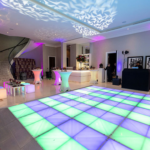 A large room featuring a huge LED dancefloor showing green and purple coloured LED panels. LED poseur tables sit around the edges, a DJ booth to one side. An ornate staircase winds upwards to one side. Lighting effects are projected onto the ceiling